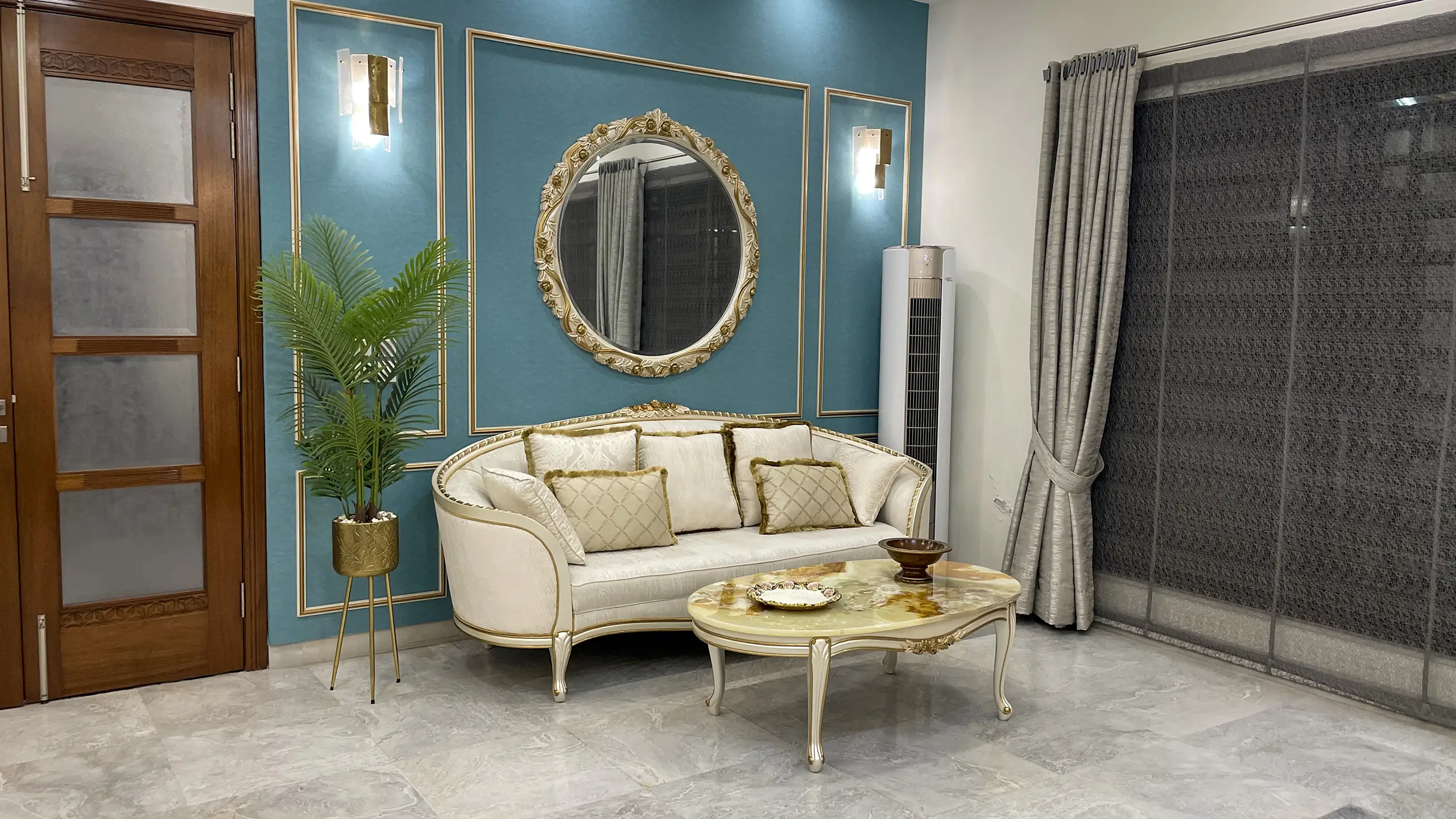 Classic living room with a curved off-white sofa, marble-top coffee table, and an ornate gold mirror on a teal wall.